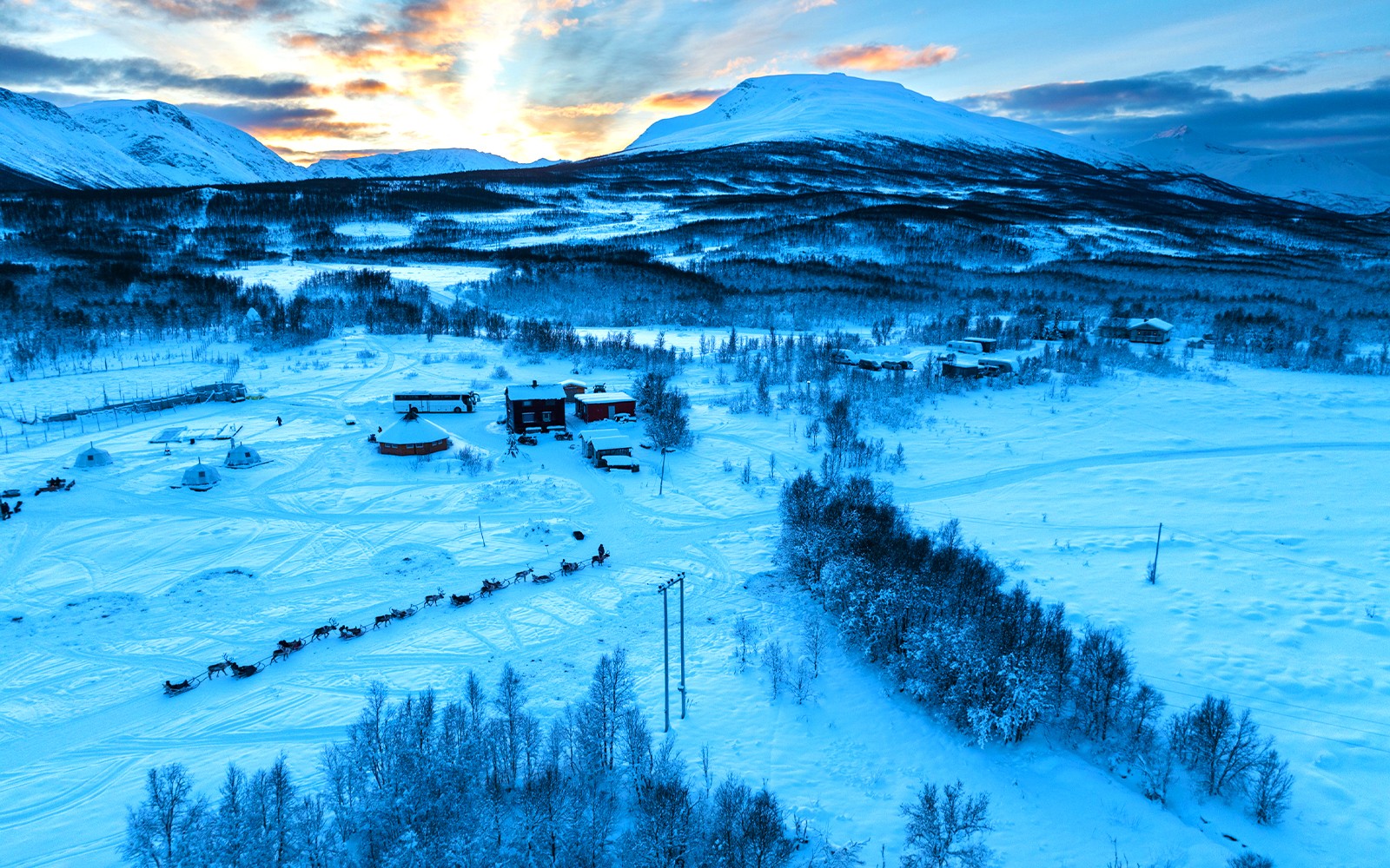 Reindeer sledding through snowy landscape with Sami tents and mountains in the background.