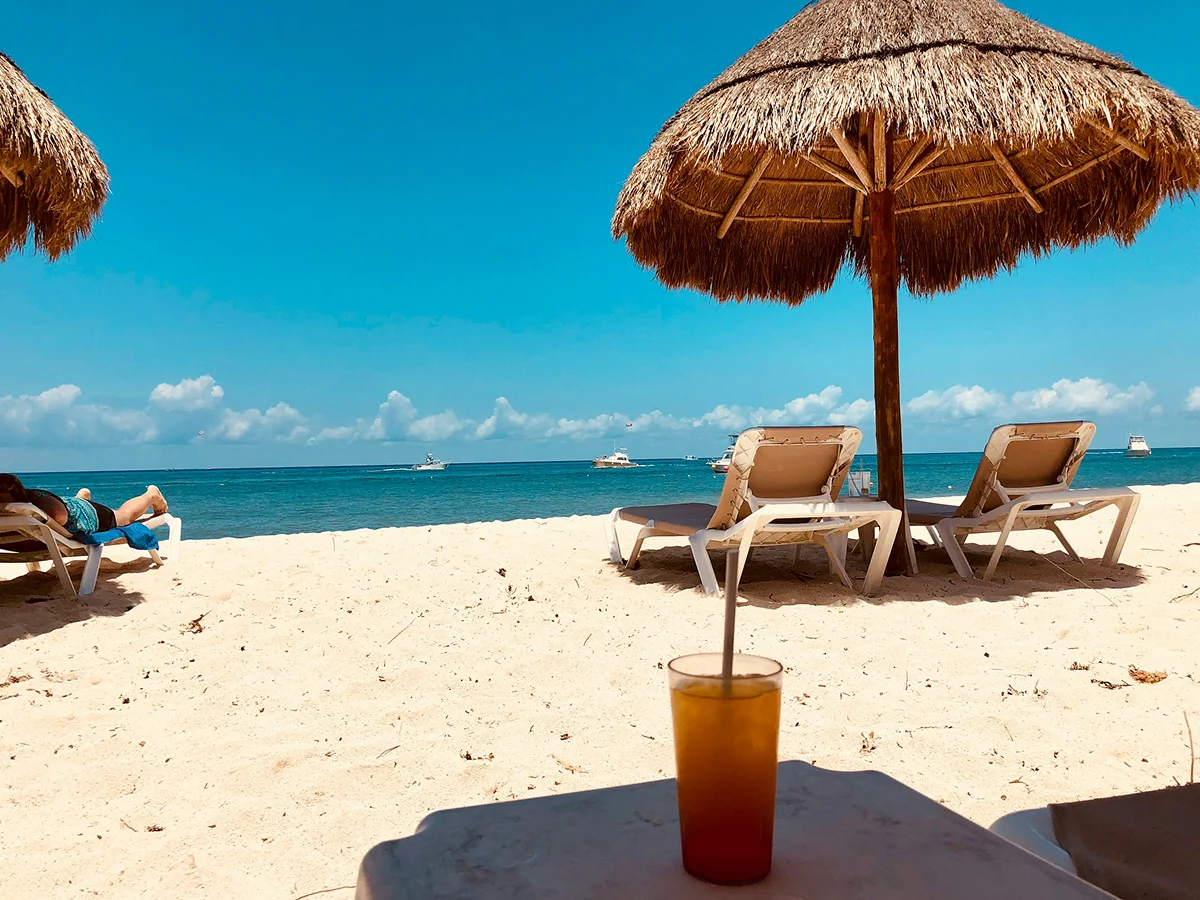 Beach lounge chair under a thatched umbrella with a cold drink and ocean view.