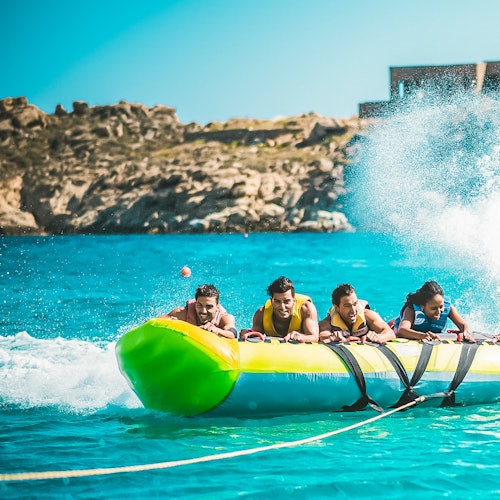 Four people ride an inflatable banana boat over turquoise water, with waves splashing, while rocky cliffs are visible in the background.