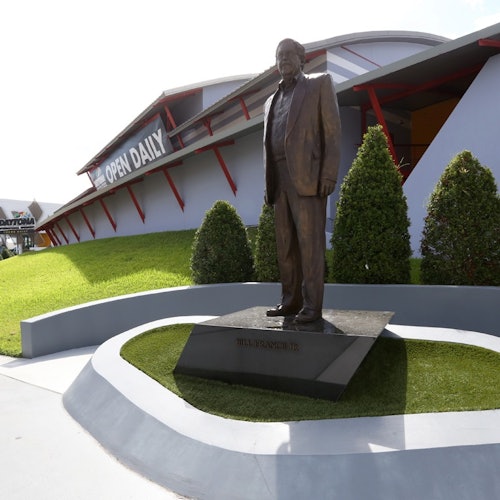 Bronzestatue af en mand i jakkesæt på en piedestal foran en bygning med et skilt, der lyder "ÅBEN DAGLIGT," omgivet af buske.