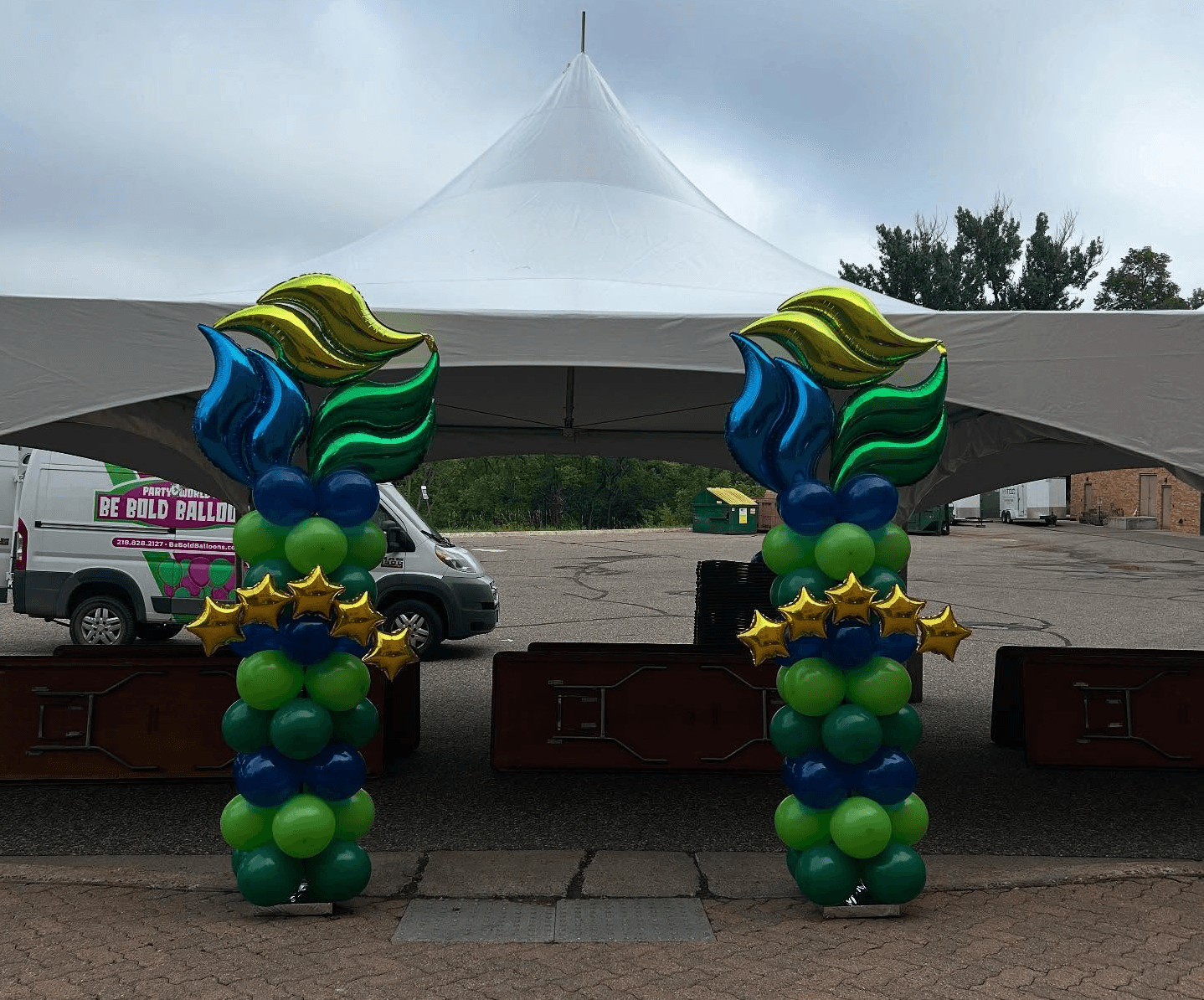 Pair of blue, green, and yellow balloon columns with starburst toppers displayed outdoors by Be Bold Balloons