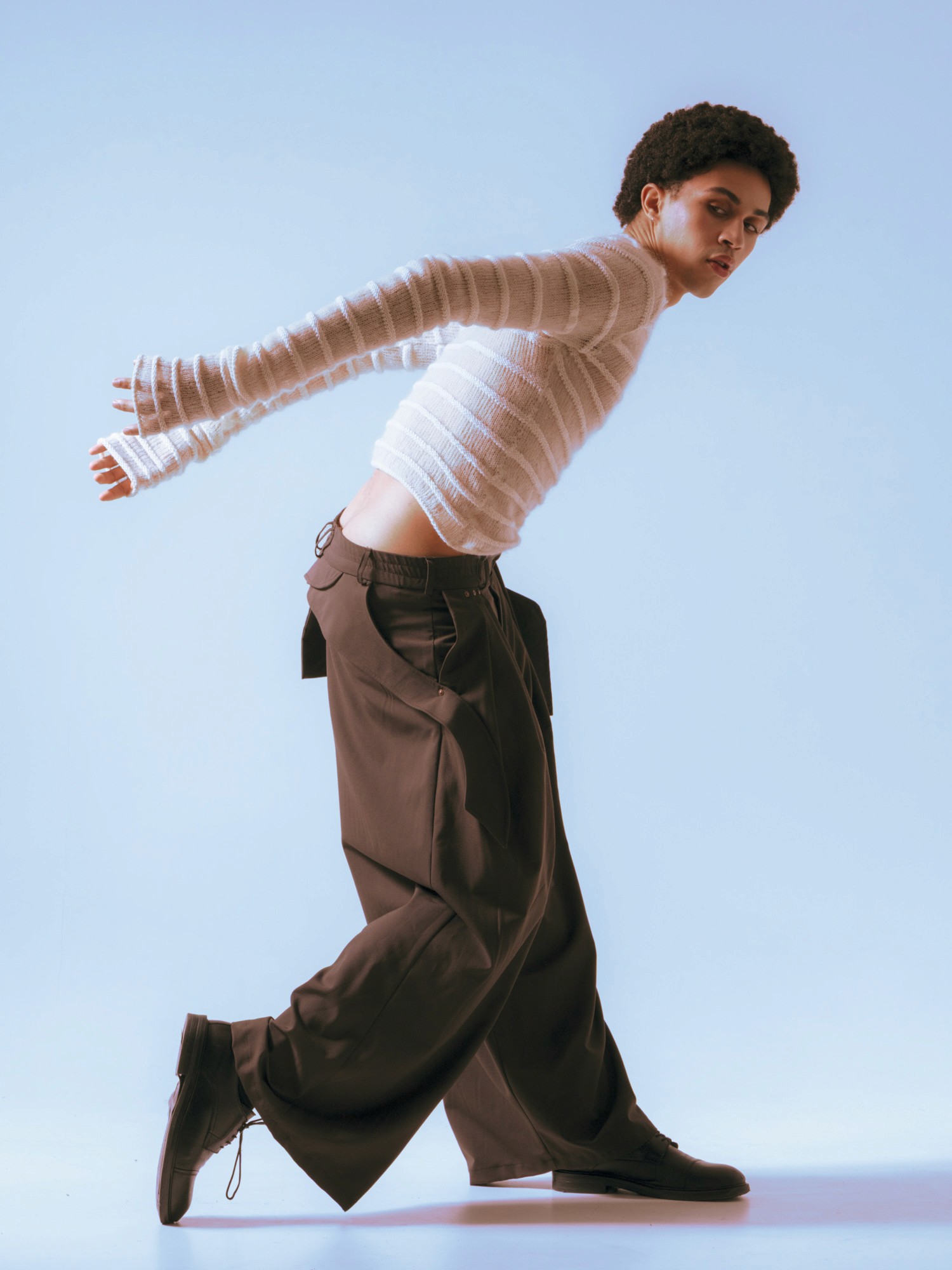 A person in a white, textured sweater and loose-fitting dark pants strikes a dynamic dance pose against a soft blue background, conveying a sense of movement and style.