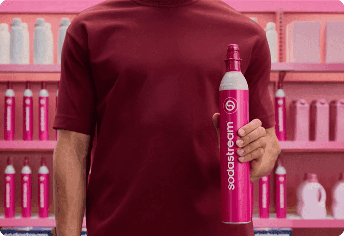 Kéz rózsaszín Sodastream palackot tart, háttérben sorba rendezett palackok.