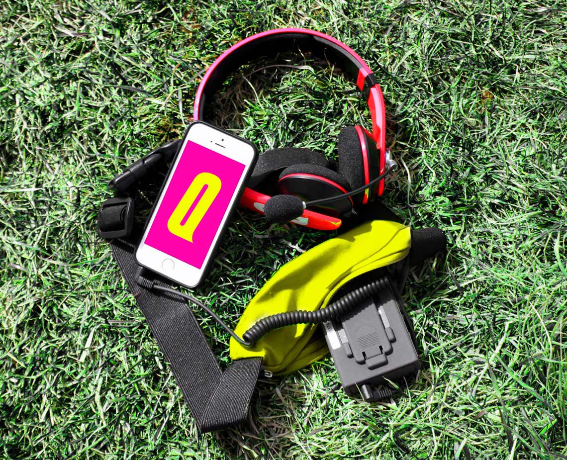 A smartphone displaying a pink screen with a yellow "0" is placed on a grassy surface. A pair of red and black over-ear headphones rests beside it. A black cable with a yellow connector is connected to the phone. A bright yellow cloth is partially visible near the headphones. The phone is positioned diagonally on the grass.