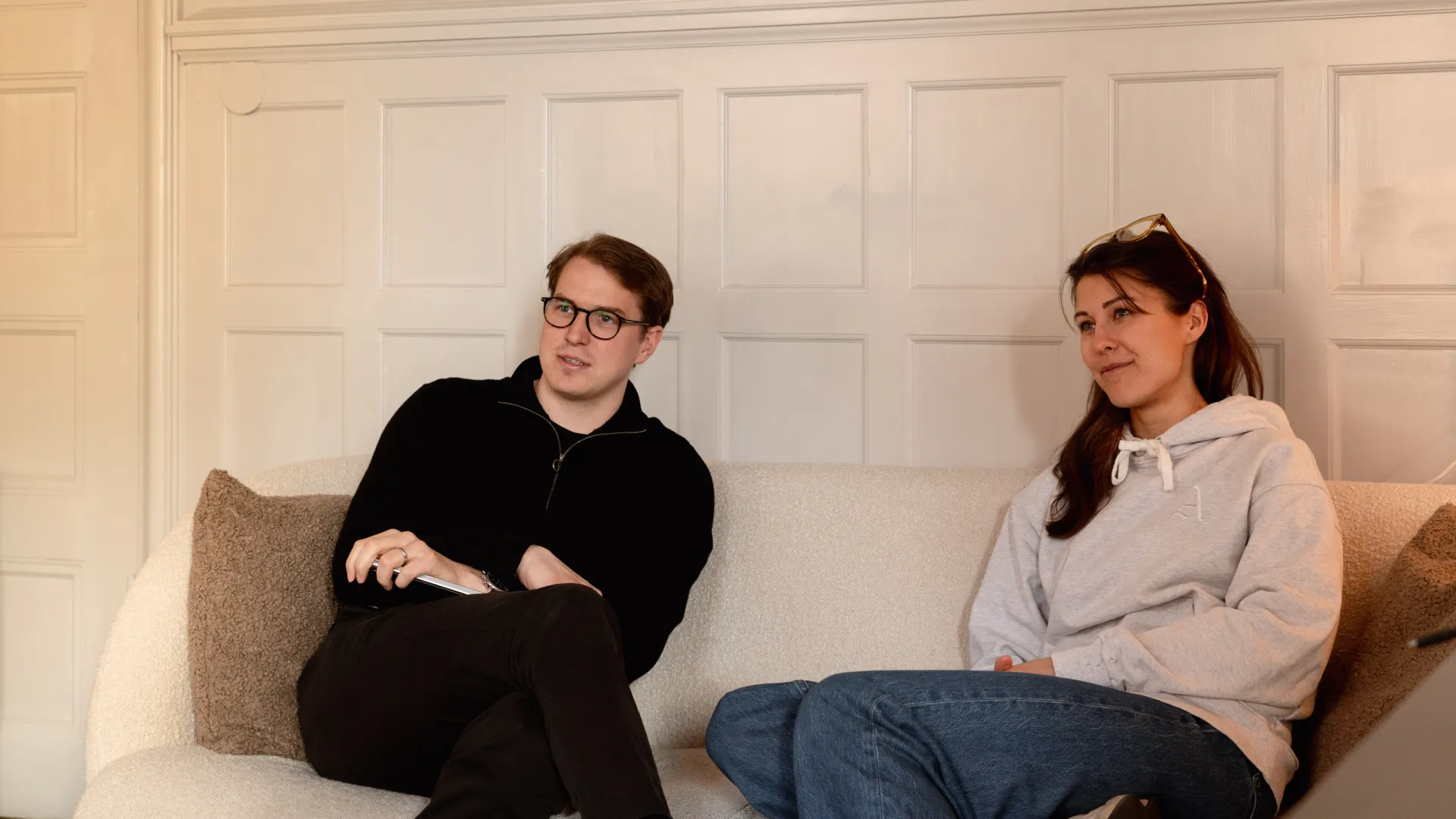 wo people sitting on a beige couch, one wearing glasses and holding a notepad, the other looking relaxed in a hoodie.