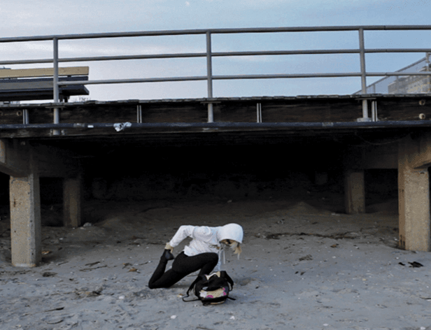 A person in a white hooded sweatshirt and black pants kneels on a sandy beach, leaning forward with their head down. A black backpack rests on the ground beside them. A concrete structure with a metal railing is visible in the background. The sky is light blue with faint clouds.