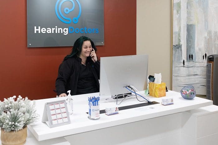 A woman sits at a reception desk, talking on the phone, with a computer and decorative items around her.