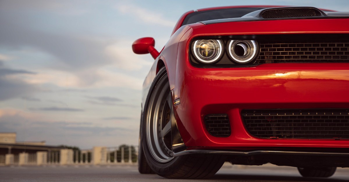 A ground-level view of the front of a red sports car. The vehicle is parked in an empty area with buildings in the distance.