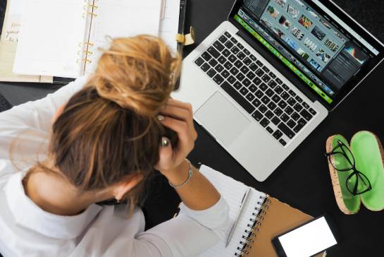 A person with a topknot hairstyle sits at a desk cluttered with an open laptop displaying photo editing software, surrounded by a notebook, smartphone, glasses, and various papers, suggesting a busy work environment.