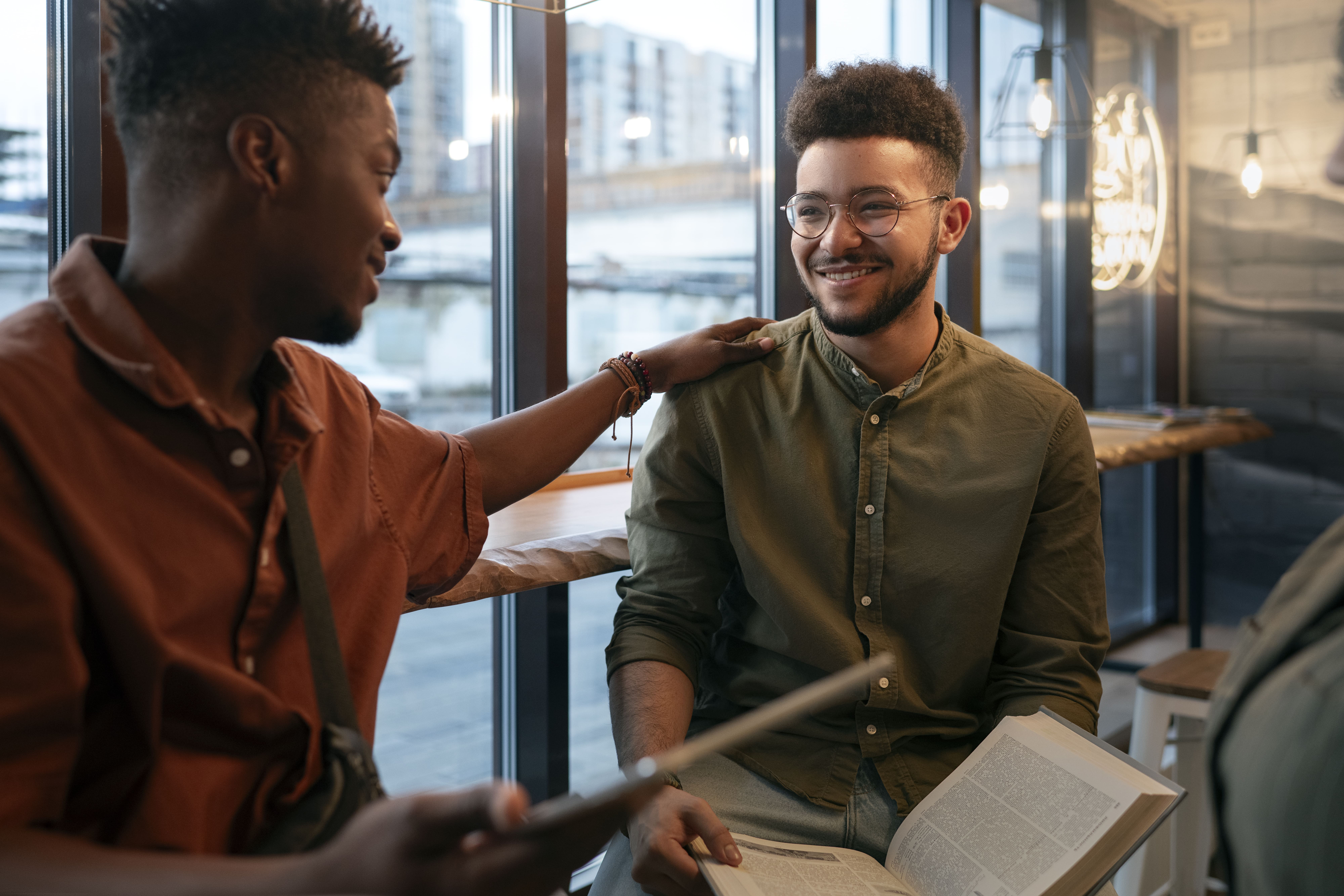 A imagem retrata dois amigos conversando e interagindo de maneira amigável em um ambiente aconchegante, provavelmente um café ou espaço social. À esquerda, um homem vestindo uma camisa marrom toca o ombro do amigo, que está à direita, sorrindo e vestindo uma camisa verde-oliva. O amigo que sorri segura um livro aberto no colo, sugerindo que estavam discutindo algo interessante. Ao fundo, grandes janelas mostram um ambiente urbano, e luzes pendentes contribuem para a atmosfera acolhedora e casual do local.