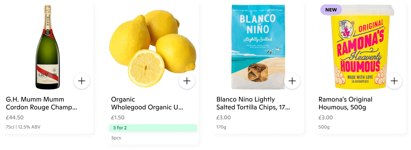 Four digital product listing cards on a white background:  G.H. Mumm Cordon Rouge Champagne: A 75cl bottle, 12.5% ABV, priced at £44.50.  Organic Wholegood Lemons: A 3-piece pack priced at £1.50, featuring a green "3 for 2" promotional badge.  Blanco Nino Tortilla Chips: A 170g bag of lightly salted white corn tortilla chips priced at £3.00.  Ramona's Original Heavenly Houmous: A 500g tub priced at £3.00, marked with a purple "NEW" badge.