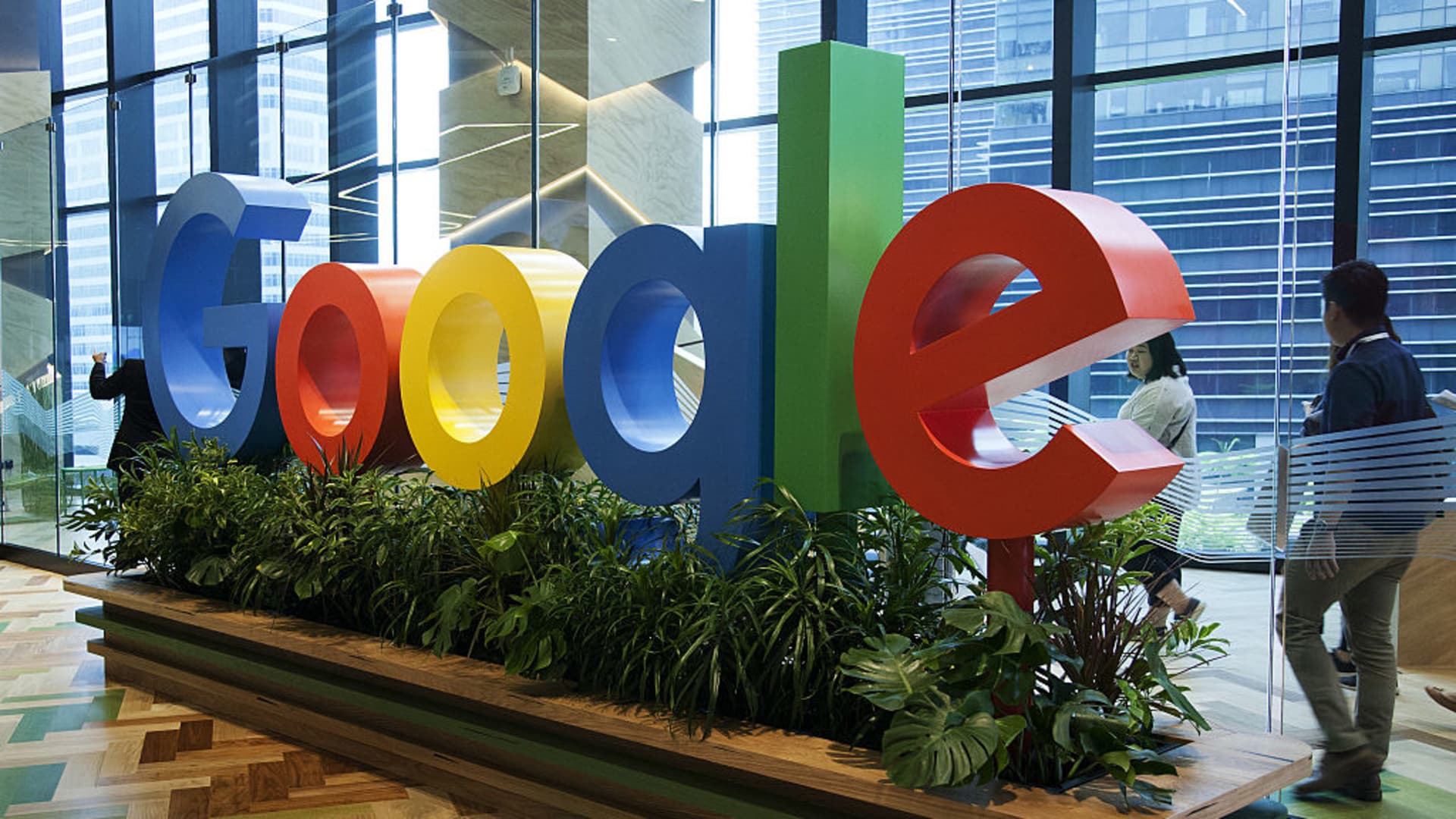 Colorful Google logo in blue, red, yellow, and green, prominently featured on a flowery platform, surrounded by people walking around.