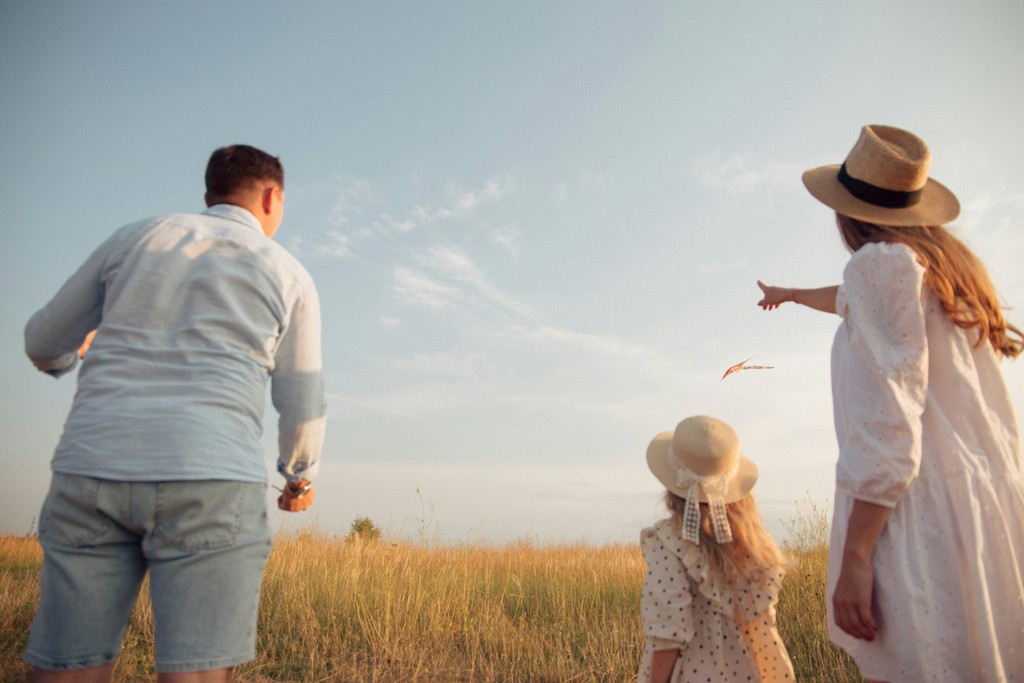 A family of three enjoys a sunny day in a meadow, flying a kite. The parents and their young daughter, all dressed in light summer clothing and hats, look towards the kite soaring in the sky. The image captures the essence of family bonding, outdoor fun, and the simple joys of spending time together in nature.