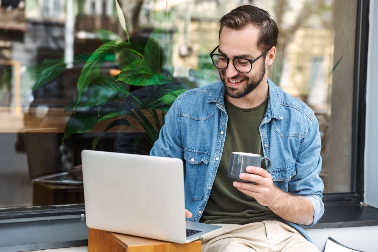 A smiling man in glasses sits at a table, working on a laptop and holding a coffee cup in a bright cafe setting.