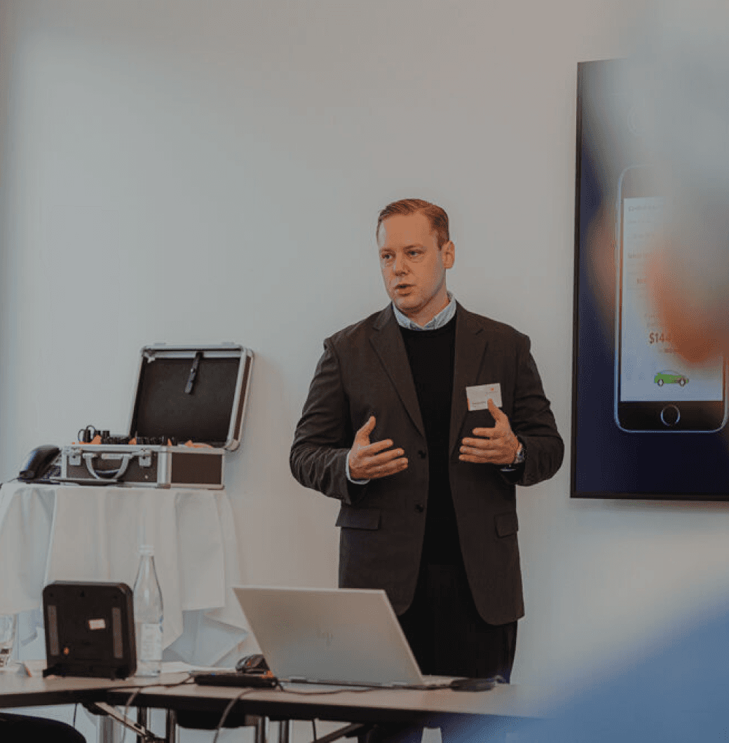A man in a suit stands by a table, gesturing while presenting in a conference room setting.