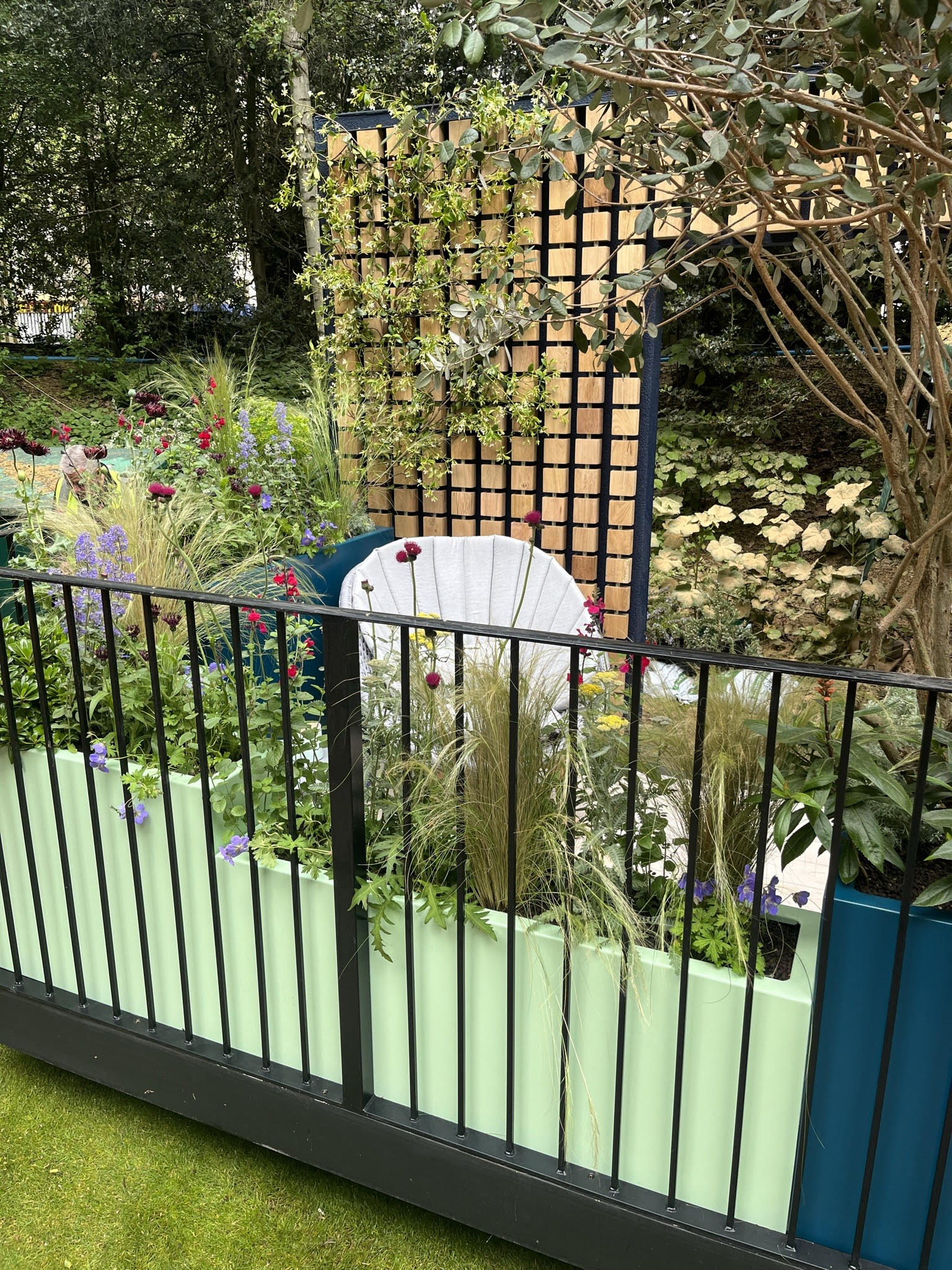 A garden view behind a black fence, featuring greenery and a small white structure in the background.