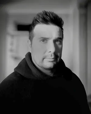 Casual studio portrait of a bearded man with curly hair in a white shirt, arms crossed, on a light gray background.