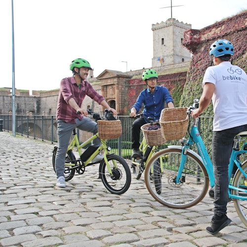 Tri biciklista s kacigama razgovaraju na popločanom putu pokraj povijesne zgrade sa zidovima prekrivenim bršljanom.