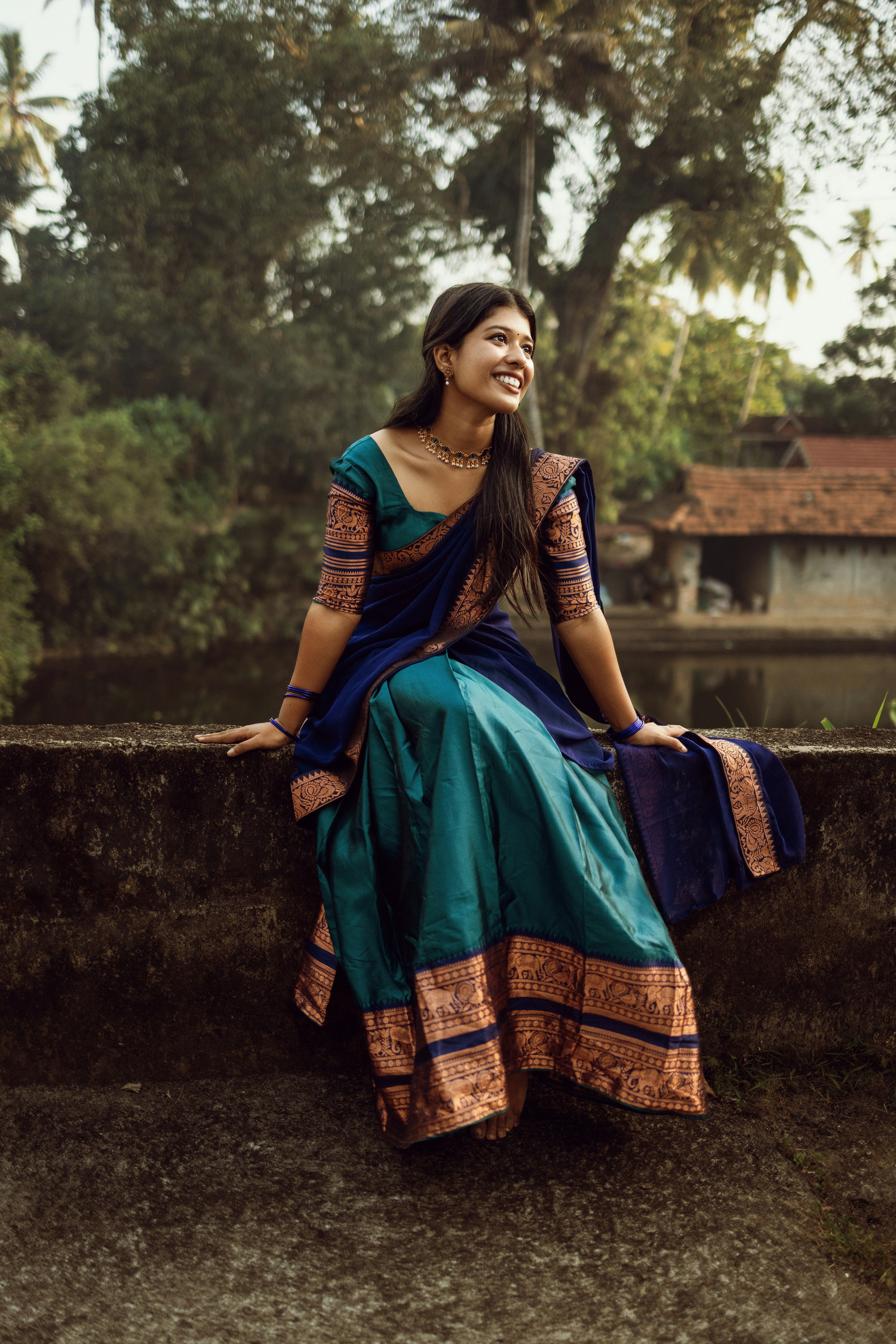 A smiling woman in a blue and teal sari sits outdoors.