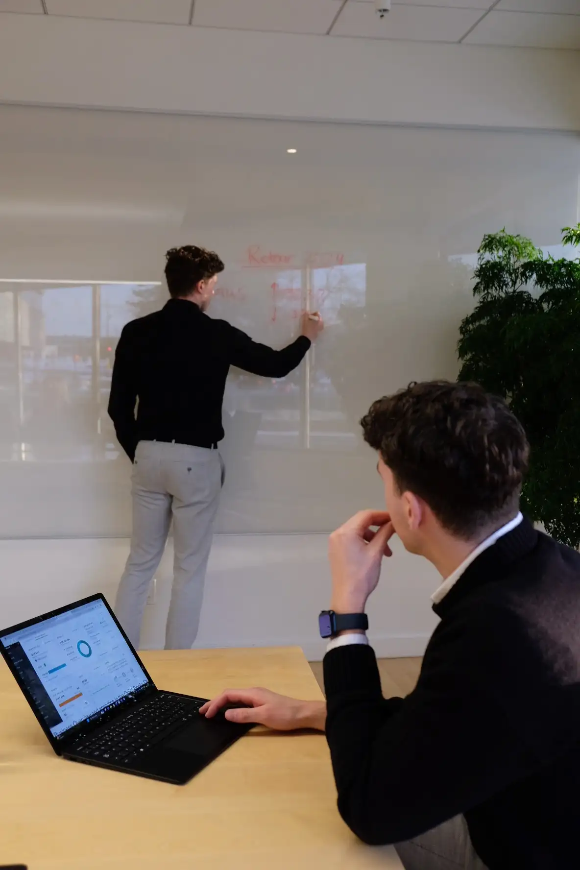 Séance de planification stratégique entre Xavier, écrivant des données financières sur un tableau blanc, et Olivier, l'observant depuis sa table de travail avec un ordinateur portable