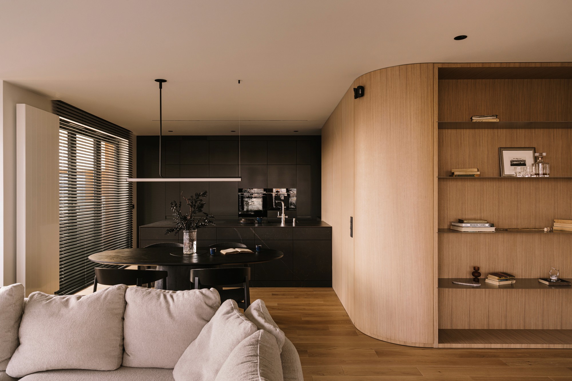 Open-plan living space with white sofa, black dining set, dark kitchen, and a curved oak shelving unit