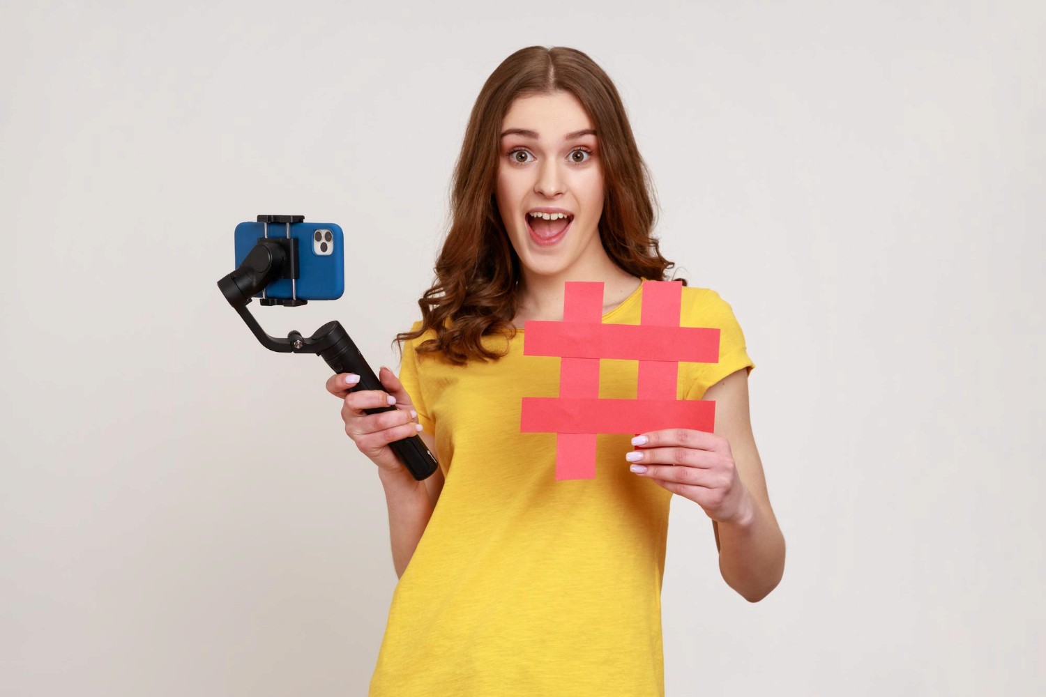 A woman smiles while holding a selfie stick and a hashtag sign, capturing a moment of joy and social media engagement.