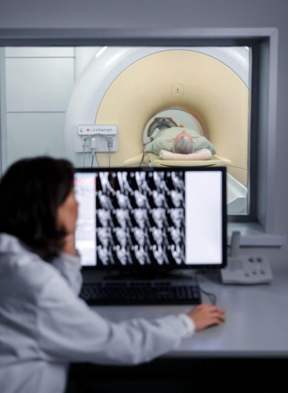 Alumna de la UCAM analizando imágenes médicas en un monitor mientras un paciente se somete a una resonancia magnética en una sala de diagnóstico hospitalario