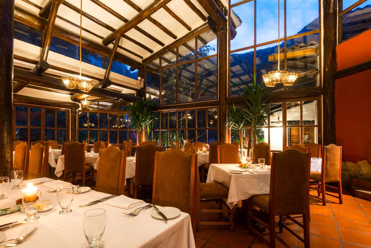 A glass-roofed conservatory restaurant with wooden beams, lit by chandeliers and candles over tables set with white linens.