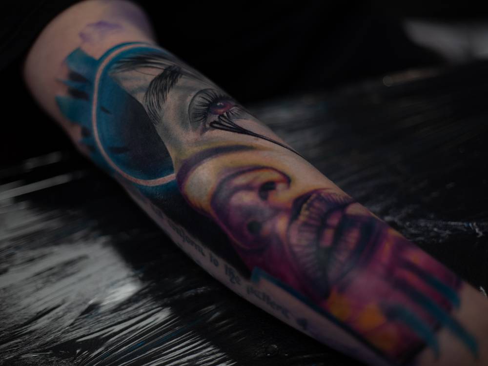 Hyper-realistic neo-noir forearm tattoo by Samma Charles, featuring a fragmented female portrait with intense shading and vibrant hues of blue, purple, and gold. The surrealist design creates a hauntingly expressive look, blending abstract elements with realism. Inked in Boston and Oxford studios