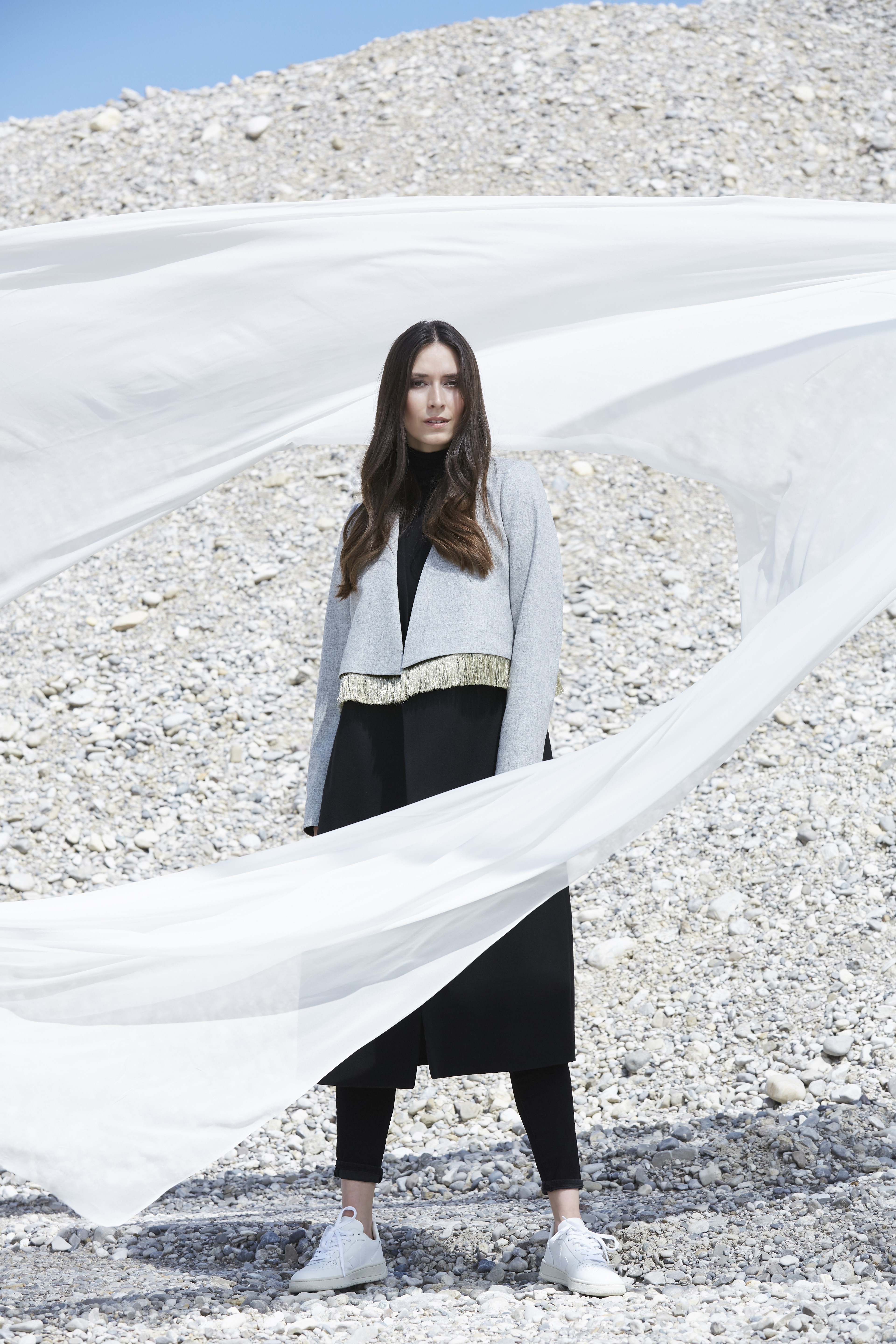 A woman stands on a rocky terrain under a clear blue sky, wearing a long gray coat and black leggings, with white fabric billowing dramatically around her in the wind.