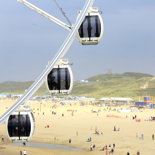 摩天輪的艙位在前景，背景中有行人正在沙灘上散步和放鬆，還有山丘和建築物。