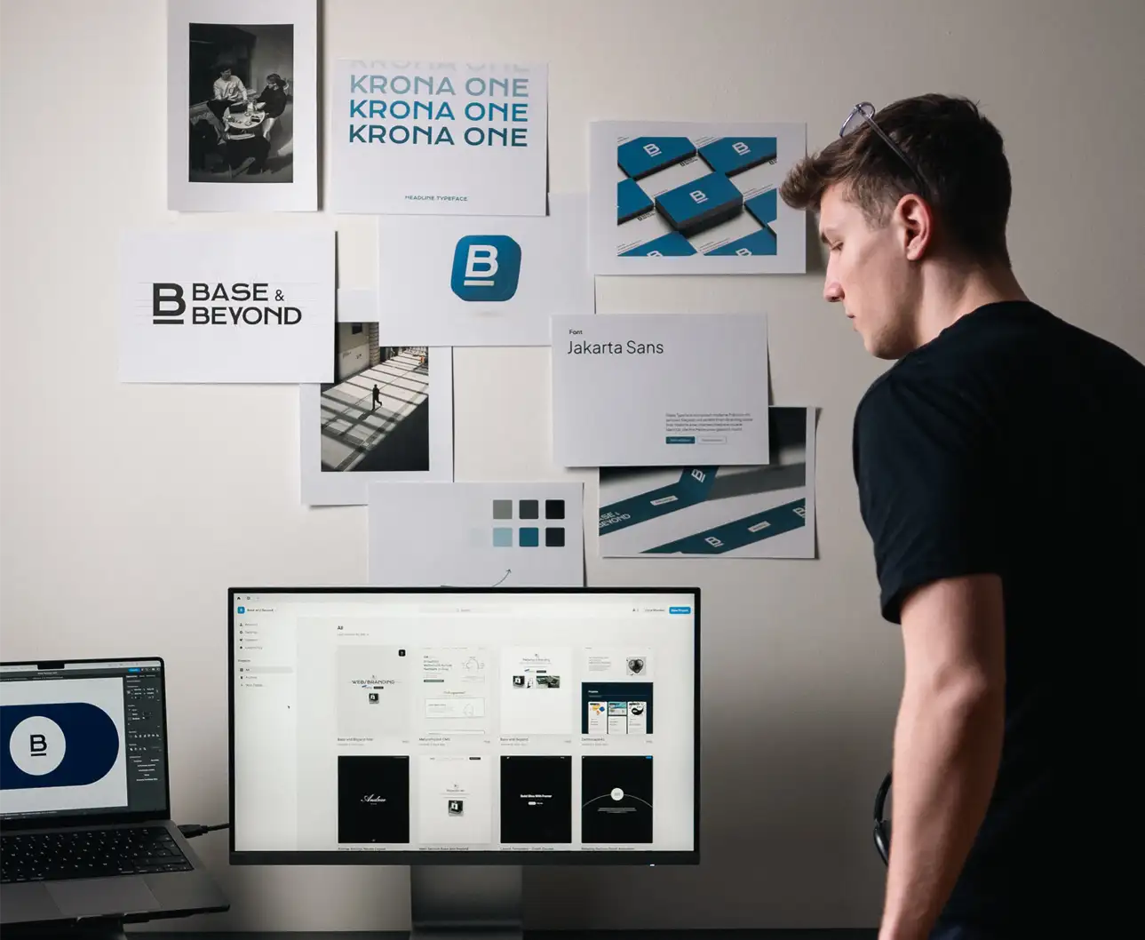 Paul Schlager arbeitet am Schreibtisch mit Branding-Materialien der Marke „Base & Beyond“ – Desktop zeigt UI-Designs, Wand dekoriert mit Logo- und Typografie-Elementen in schwarz und blau.