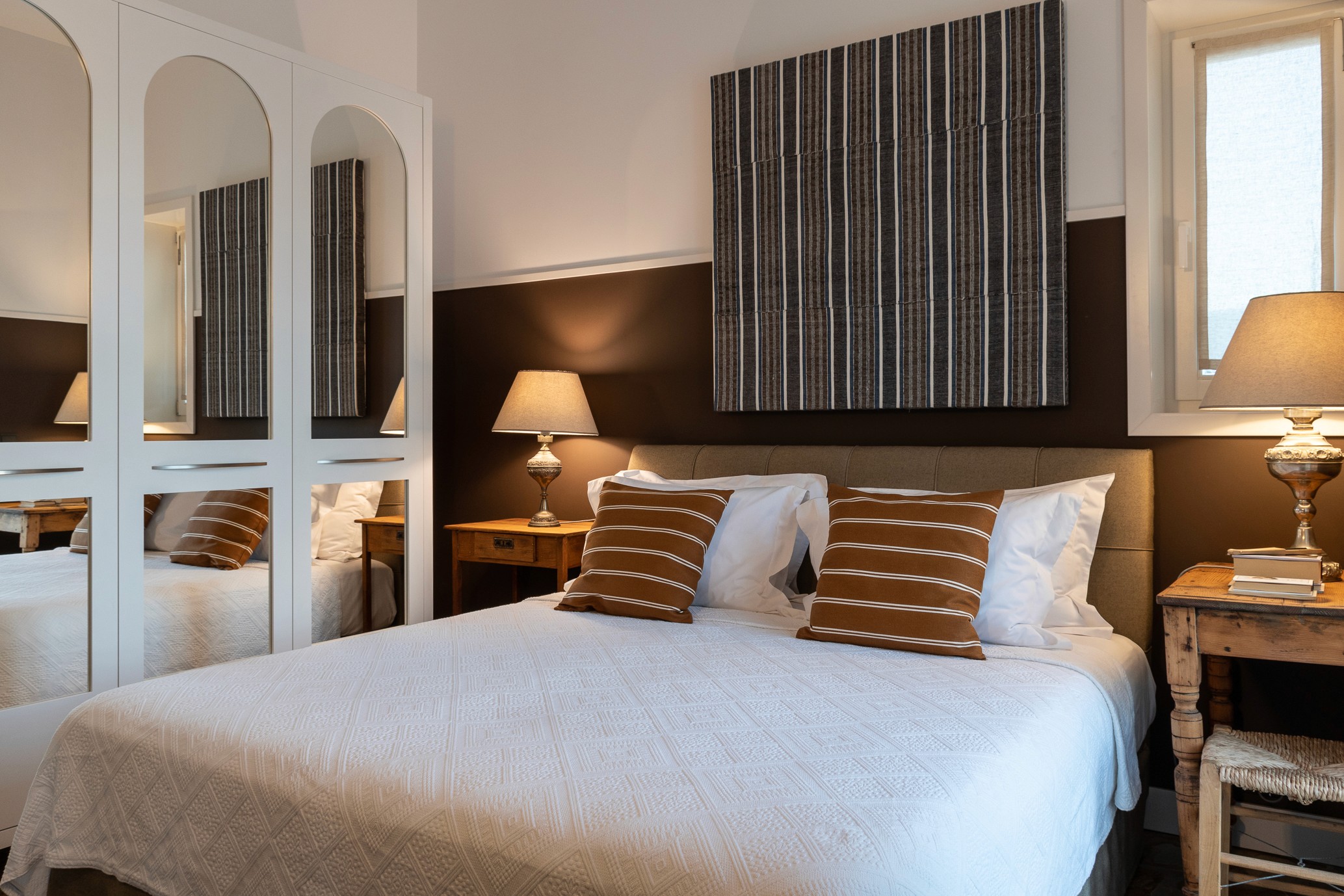 A bedroom with a bed with linen and brown and white striped pillows with an artwork hanging above - Éros Kéros