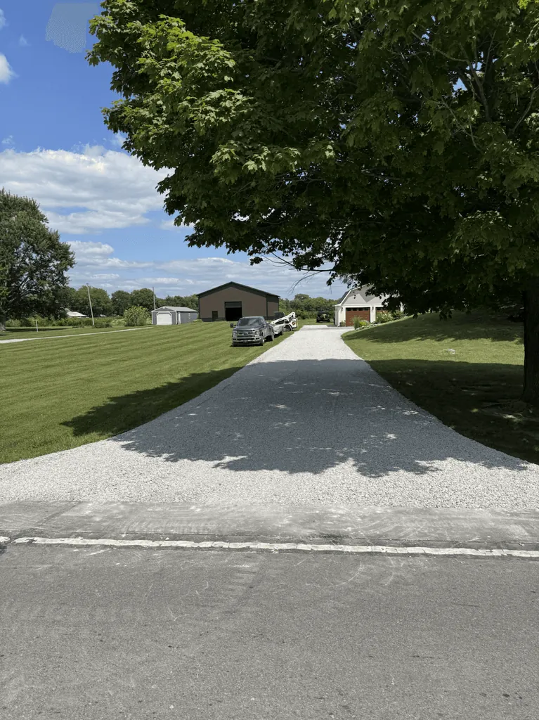 Fresh gravel driveway running from the road to a pole barn under a large shade tree