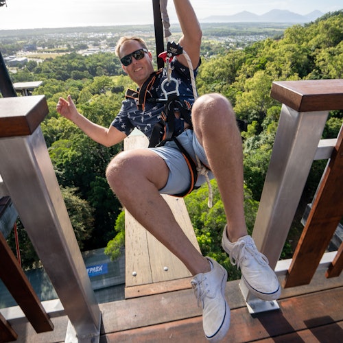 一名身穿安全带的男子准备从山林区域滑索而过，背景中可以看到远处的地平线和山脉景观。