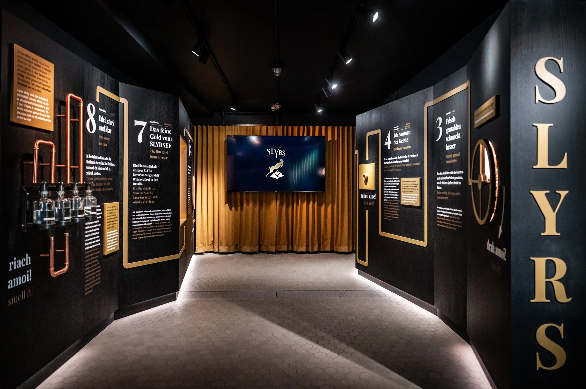 SLYRS whisky exhibition corridor with copper distillation pipes mounted on dark walls, illuminated story panels and a golden curtain framing a video screen.