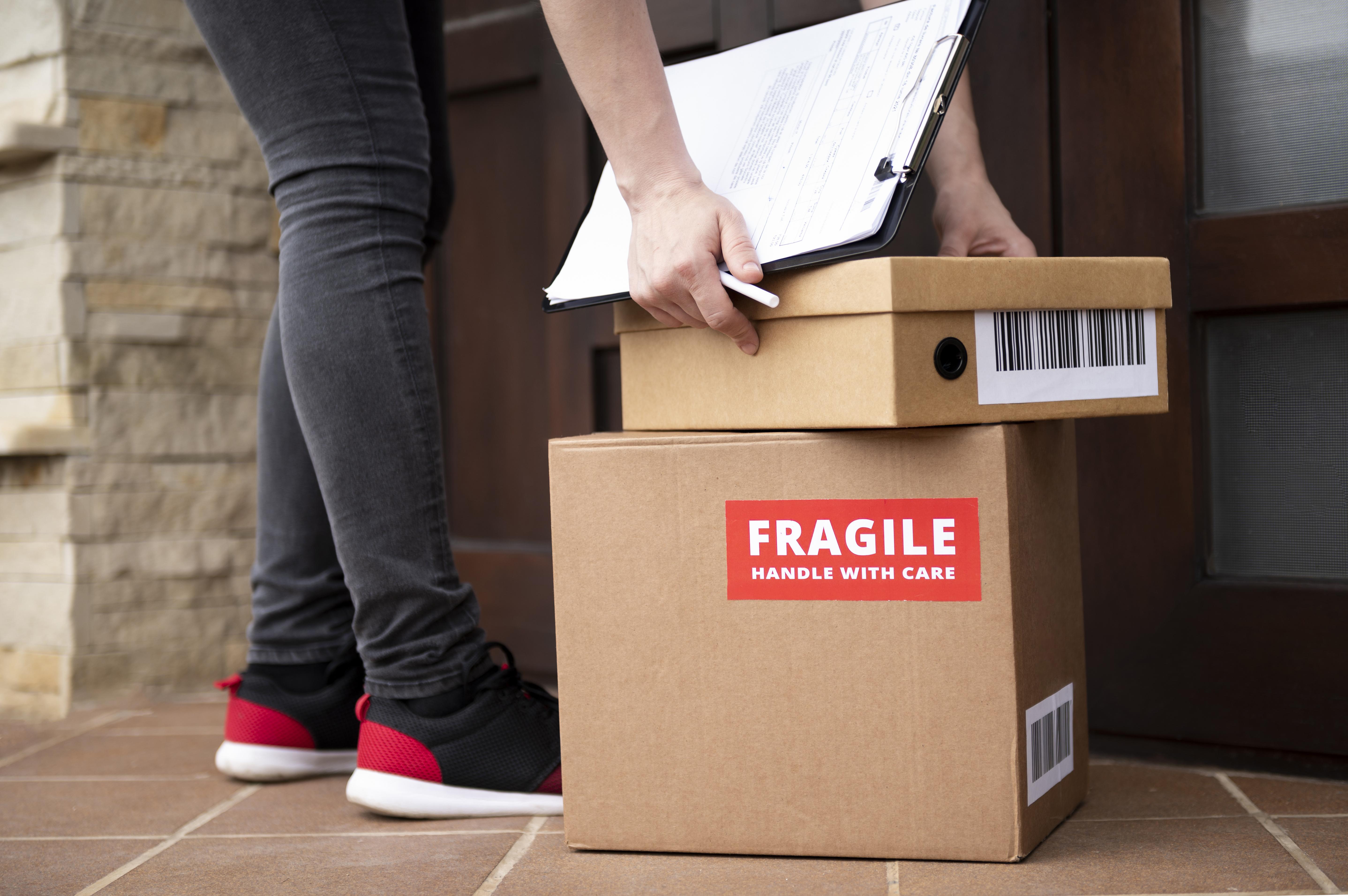 Person in red sneakers delivers a large box labeled "Fragile" and a smaller box. They hold a clipboard over the boxes at a doorstep.