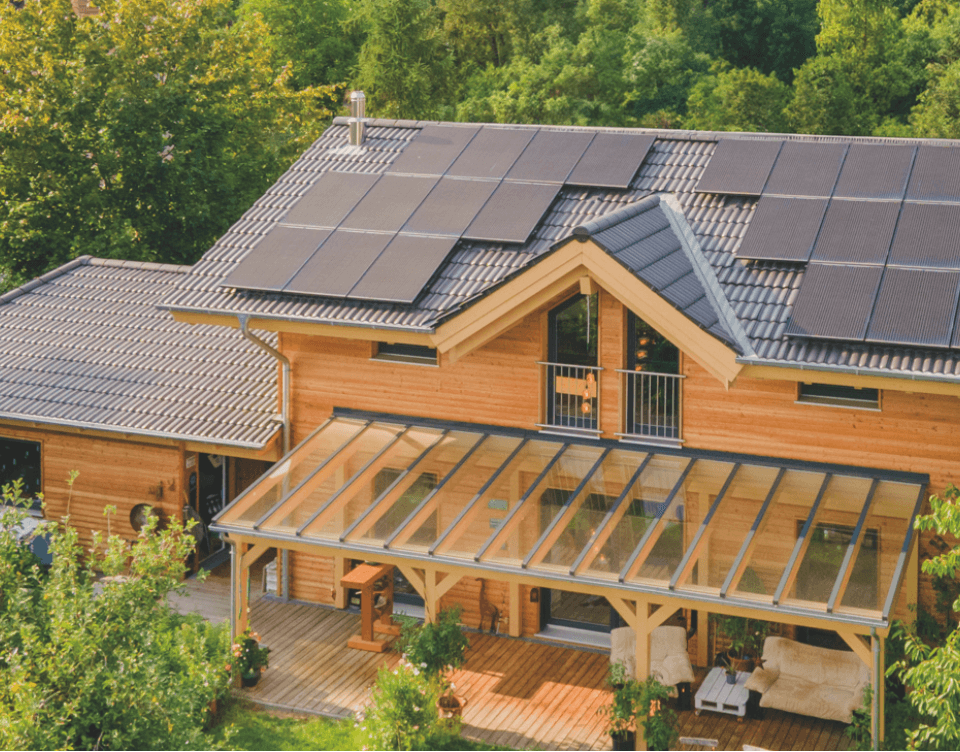 Holzhaus mit Satteldach und vielen Solarmodulen, überdachte Terrasse mit Glasdach und Gartenmöbeln.