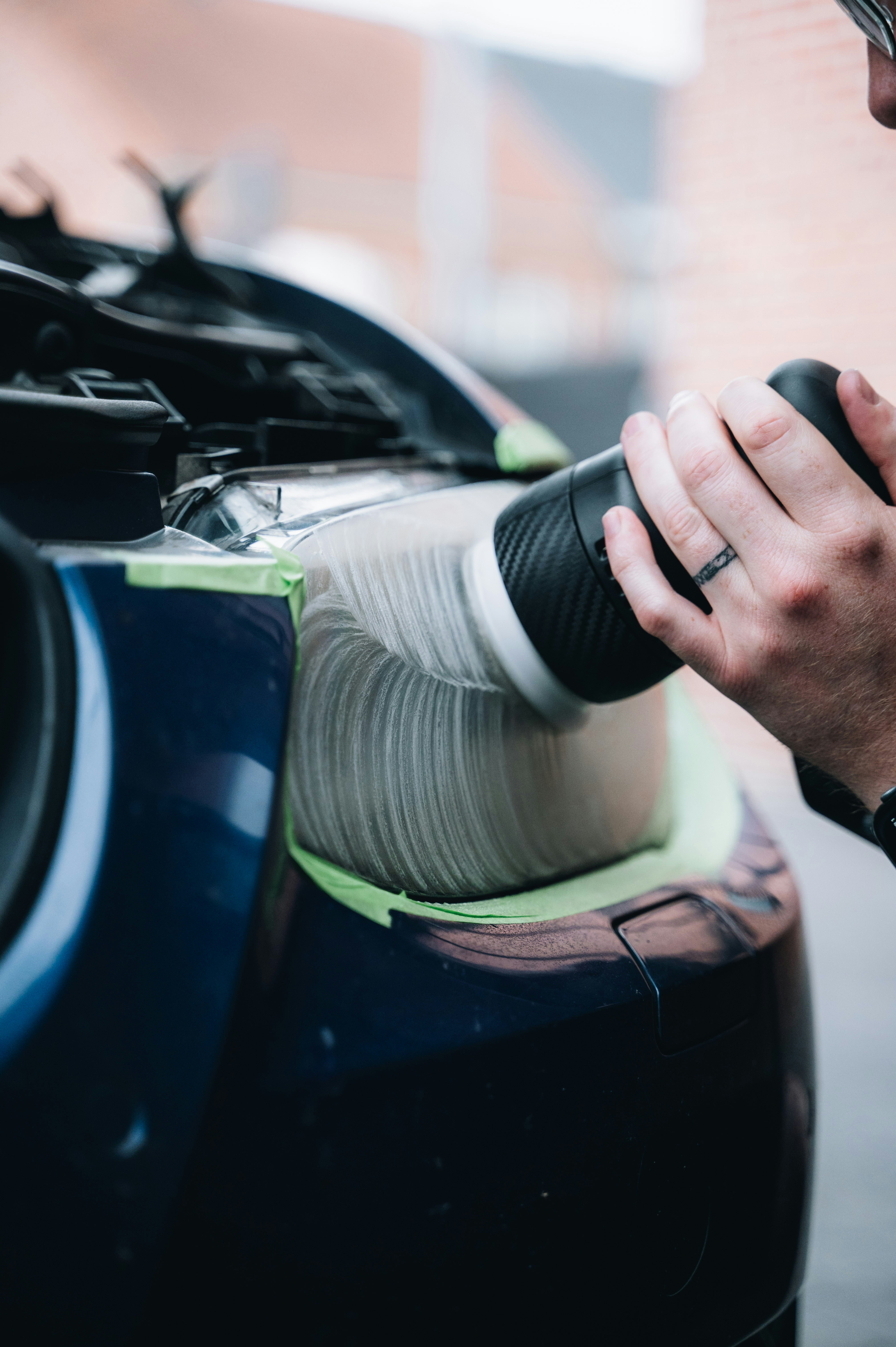 A person uses a power buffer to polish the right side of a blue car, with the area protected by green tape, in a garage setting.