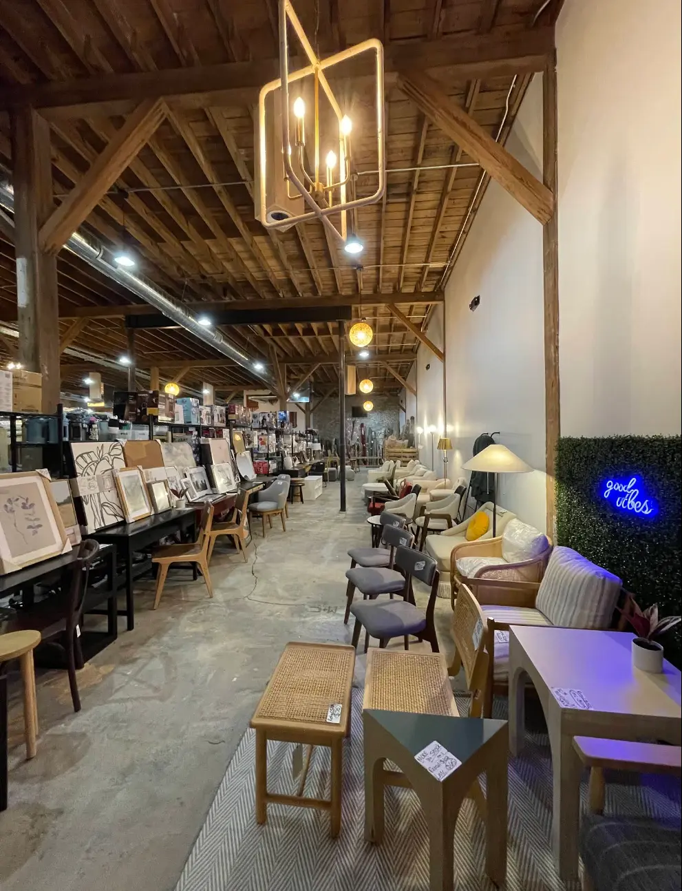 Open warehouse-style home decor store featuring furniture, artwork, and lighting displays under exposed wooden beams.
