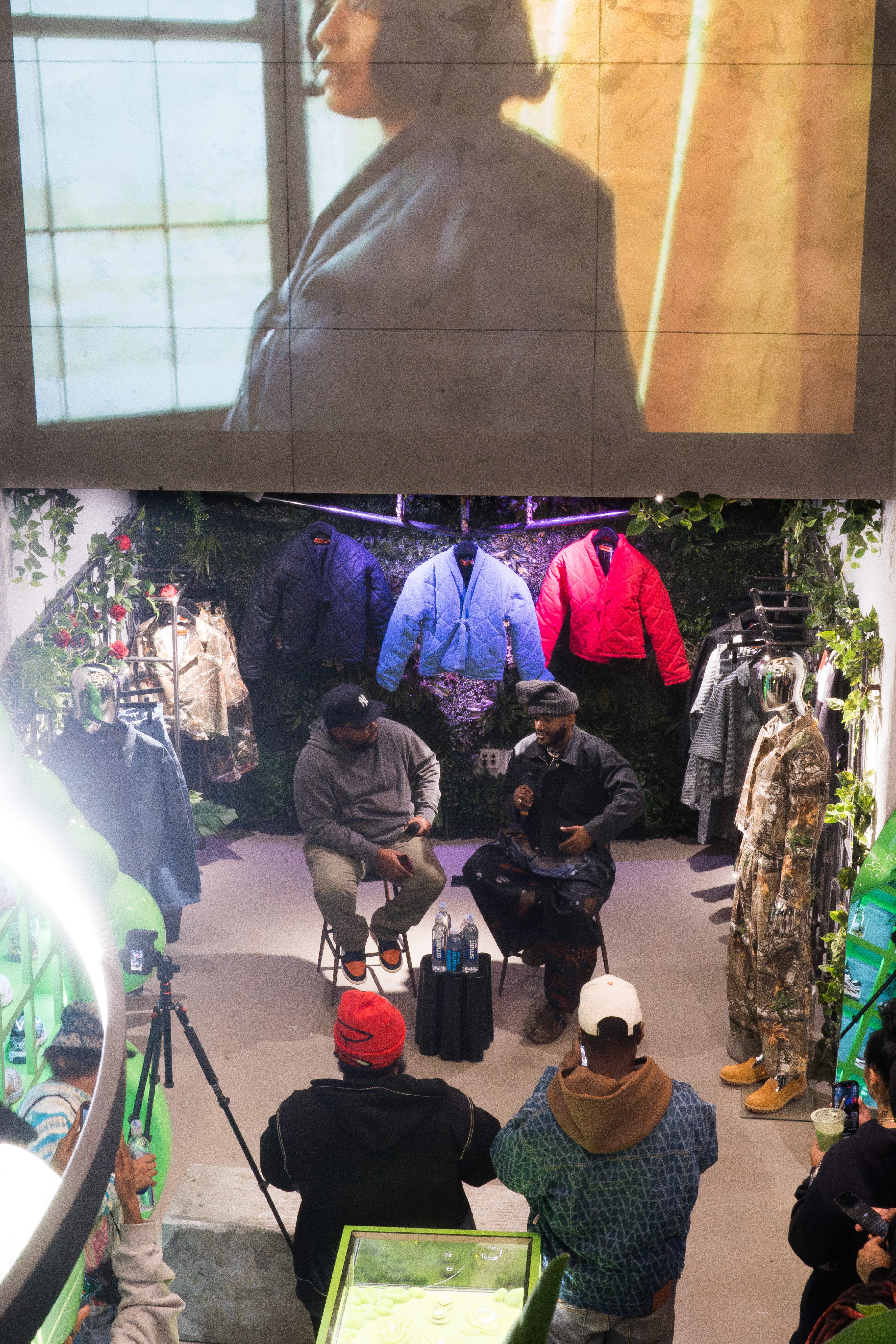 Panel conversation in a retail-style space with a clothing rack and lighting, viewed beneath a projected screen.