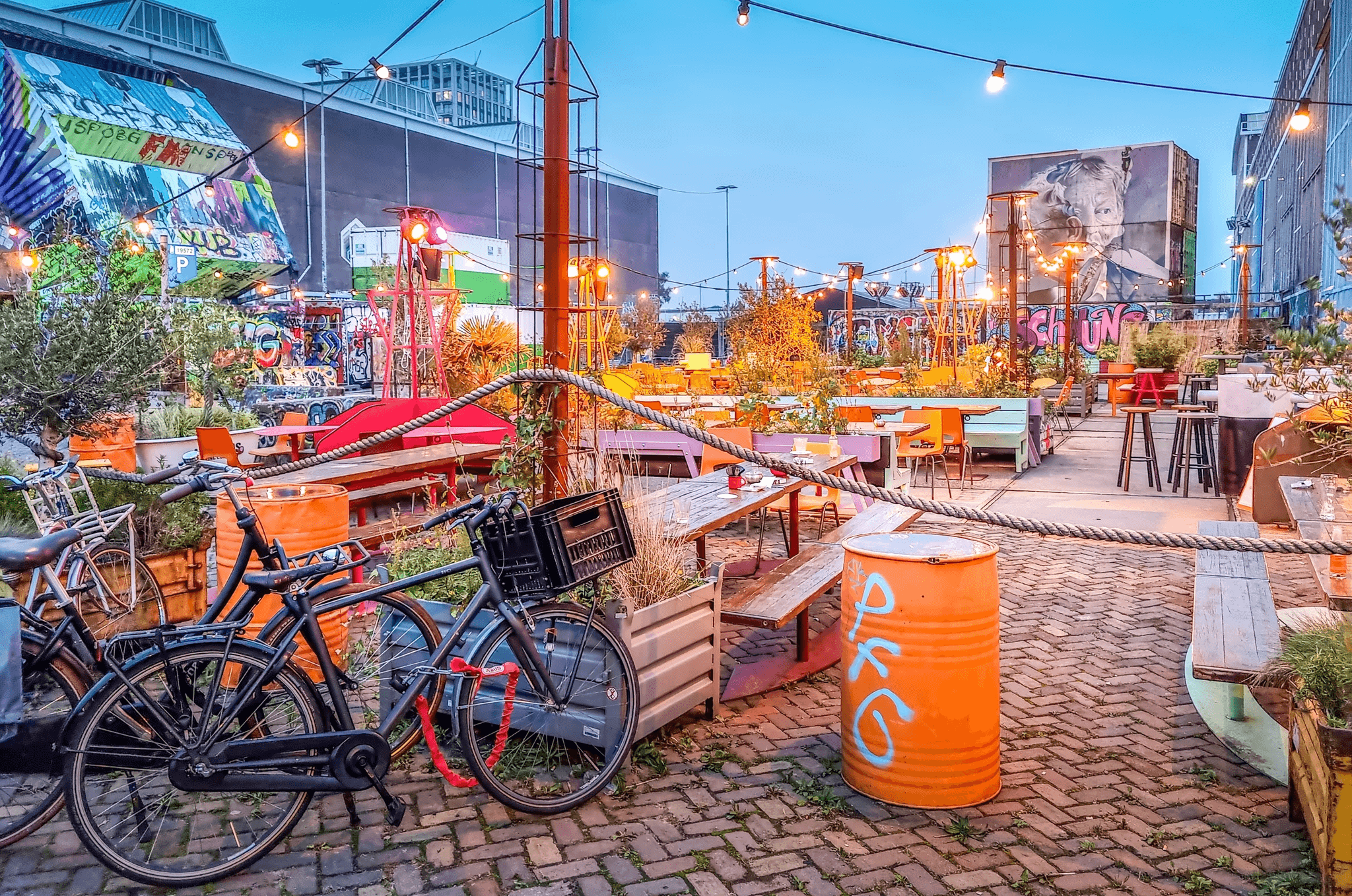 Colorful terrace in Amsterdam Noord, with bikes, string lights, graffiti-covered walls, and eclectic seating at a creative community area.