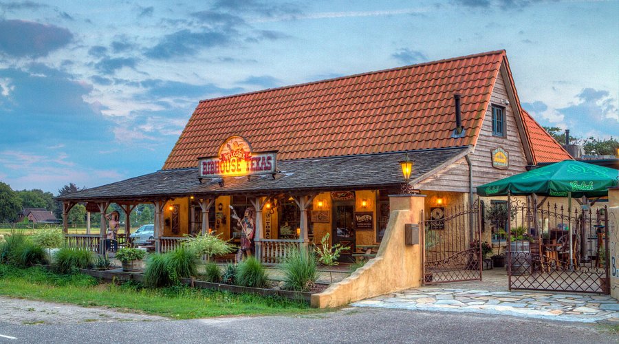 Exterieur van een restaurant met een typisch Amerikaans western-thema, genaamd Ribhouse Texas. Het gebouw heeft een rode dakpan en veranda met lantaarns.