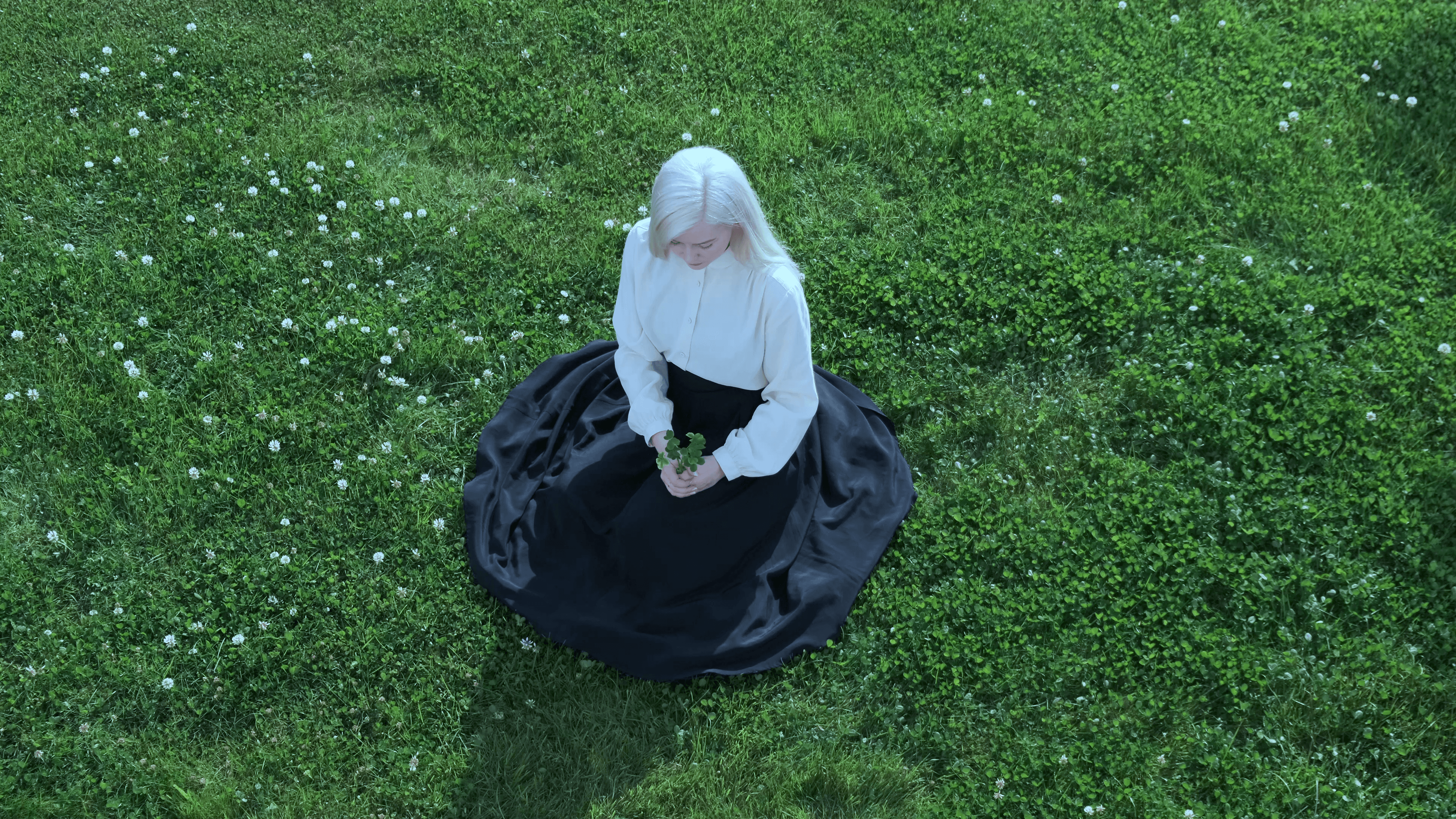 A still from Ásta Fanney Sigurðardóttir's Munnhola, obol ombla obla: viewed from above, a figure with long, pale blonde hair sits on a lawn of green clover and small white flowers, wearing a cream blouse and a wide black skirt that pools around her, holding a small sprig of leaves in her hands.