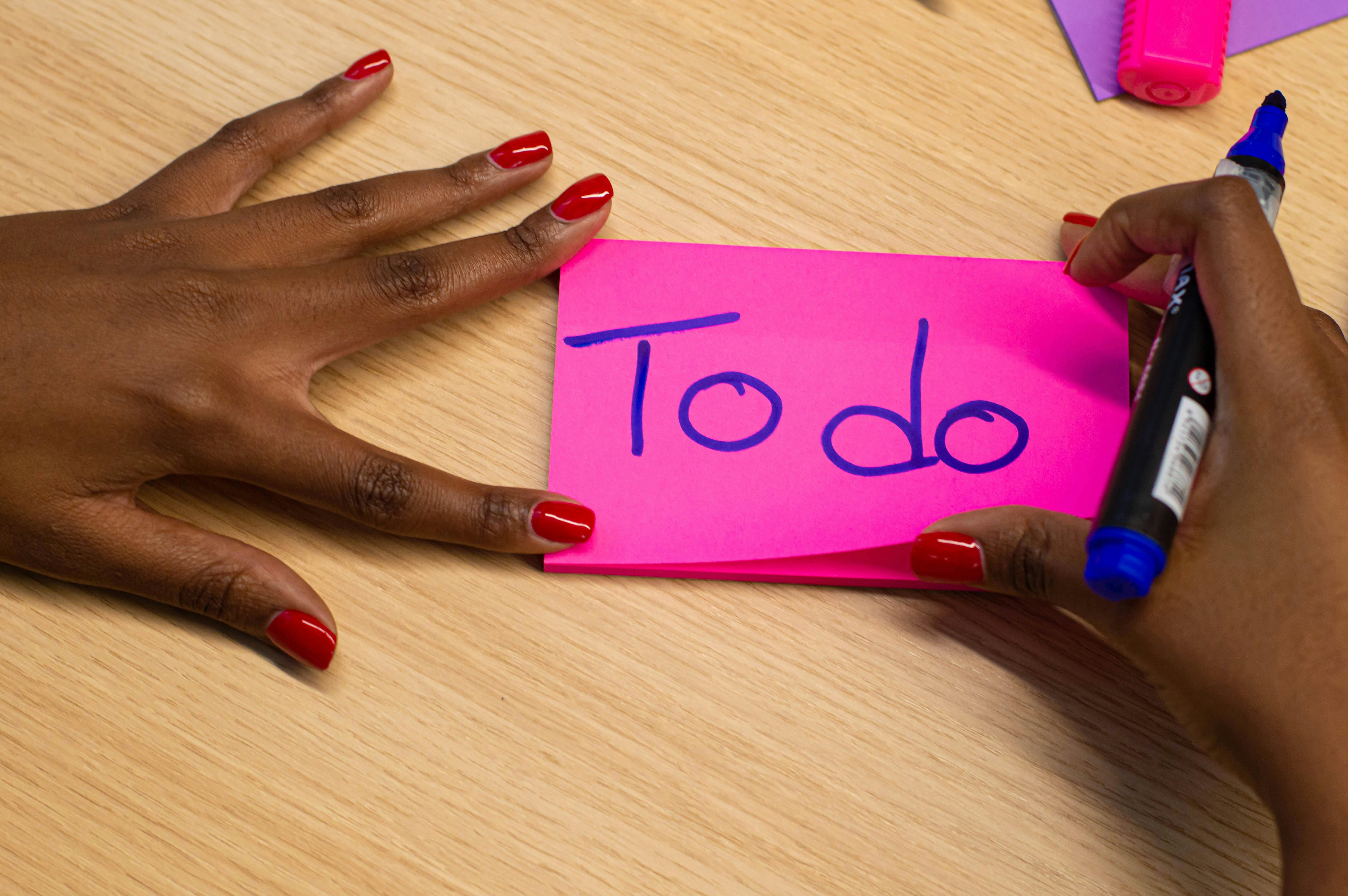 Mano femminile con smalto rosso che scrive “To do” su un post-it fucsia, simbolo di pianificazione personale e gestione delle priorità.