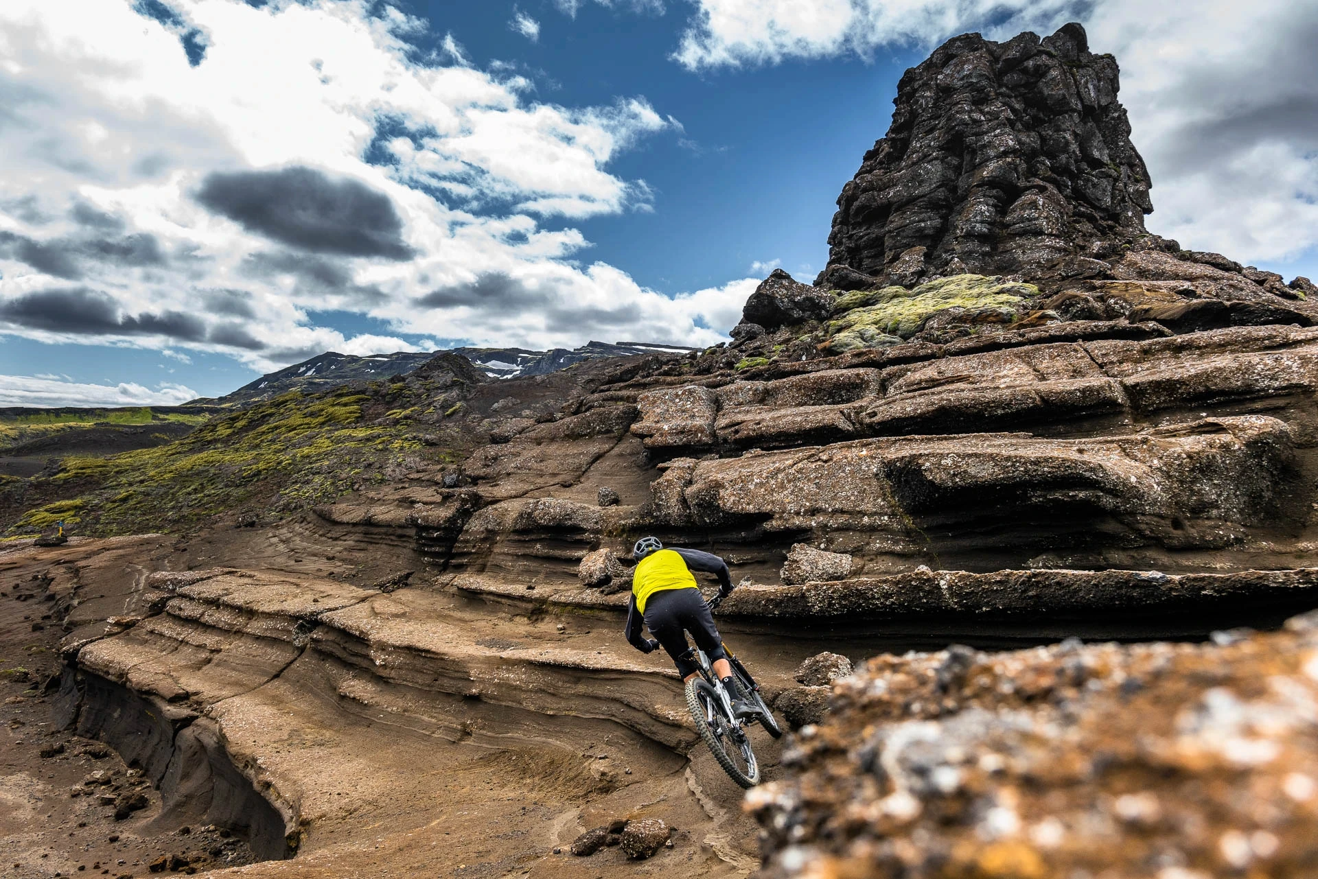 Mountain biker riding across layered rock formations in a rugged volcanic landscape.