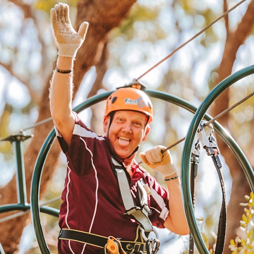 A person wearing a helmet and gloves waves while participating in a zip lining activity among trees.