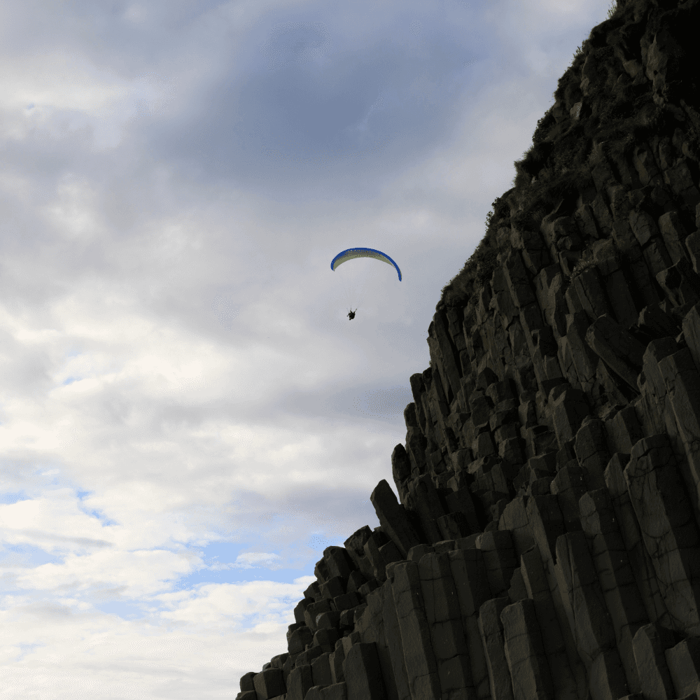 Paragliding over Reynisfjara black sand beach in Vík Iceland with basalt column cliffs and ocean below, an adventurous South Coast activity experience.