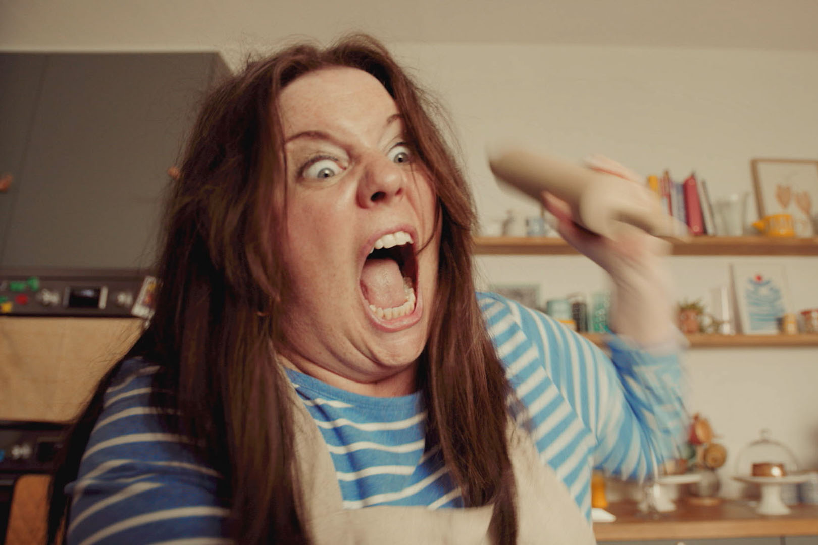 A woman with long brown hair wearing a striped blue shirt and beige apron, looking angry and screaming while holding a rolling pin in a kitchen.