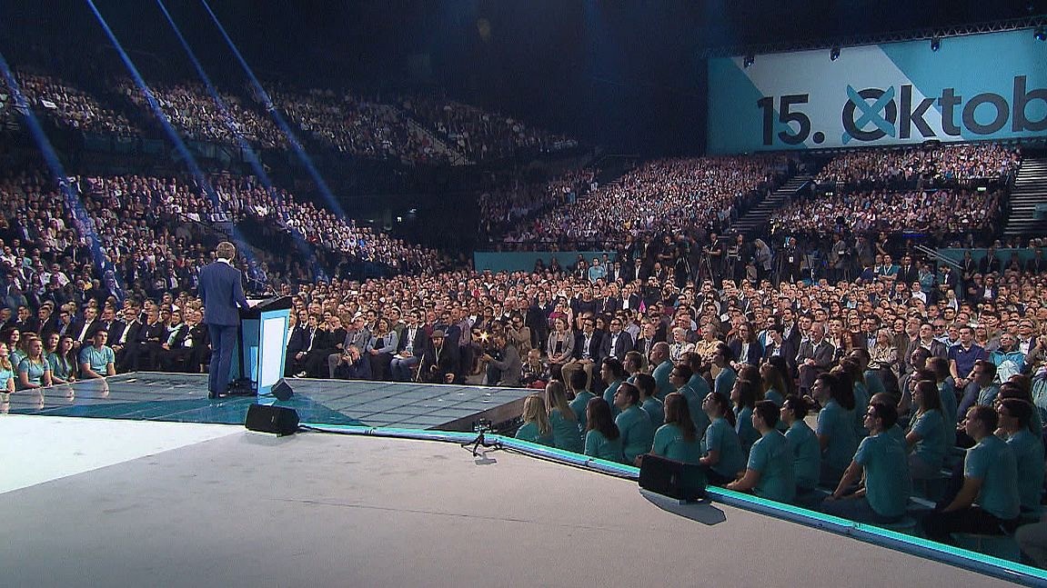A speaker addresses a large audience in a crowded venue, with a large screen visible in the background.